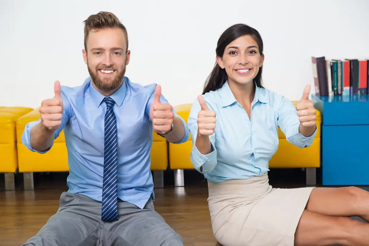 Two successful man and businesswoman showing thumbs up as sign of successfully performed work. Young managers looking at camera and sitting on floor. They working as team. Business success concept